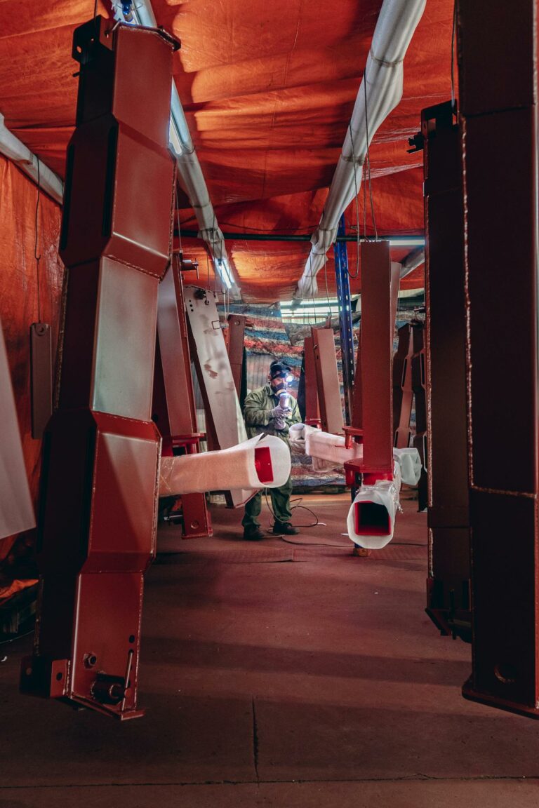 Welder working amid suspended metal structures in a factory setting.